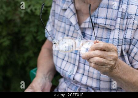 Donna anziana seduta sulla natura e tenendo gli occhiali a portata di mano, primo piano, senza volto Foto Stock