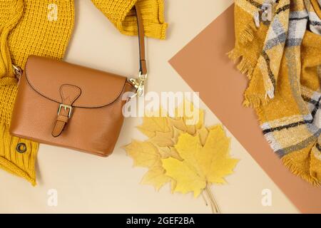 Borsa donna in pelle arancione, maglione a maglia, sciarpa, foglie autunnali su sfondo marrone beige. Accessori alla moda per l'autunno. Abbigliamento accogliente Foto Stock