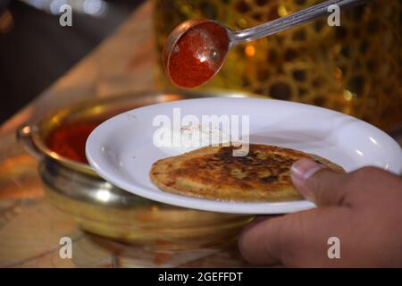 Mano di uomo indiano che porta Spice in patata di cagliata fritta nel suo piatto con un cucchiaio da una pentola grande Foto Stock
