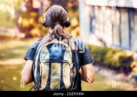 Guardando indietro adolescente scuola superiore ragazzo con cuffie e zaino cammina verso l'edificio della scuola. Concetto di ritorno a scuola. Foto Stock