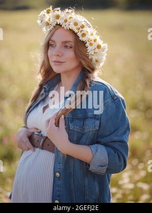 Bella donna incinta in un abito, giacca in denim e una corona di margherite rilassante fuori nel parco. Gravidanza sana e concetto di viaggio. Foto Stock