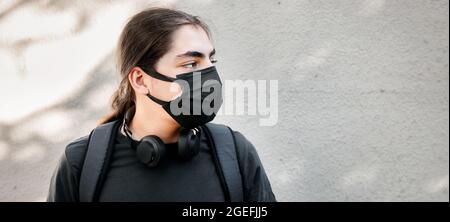 Ragazzo giovane adolescente o preadolescente con cuffie e zaino con maschera medica con sfondo bianco parete. Coronavirus pandemia e schiena Foto Stock