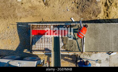 Sopra la vista dall'alto, i lavoratori edili stanno livellando il calcestruzzo fresco con il listello metallico in trincea quadrata dopo la colata dal camion miscelatore. Foto Stock