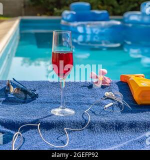 Un calice di vino rosso vicino alla piscina su un asciugamano blu con occhiali da sole, auricolari, un fiore di oleandro rosa e crema solare accanto ad esso Foto Stock
