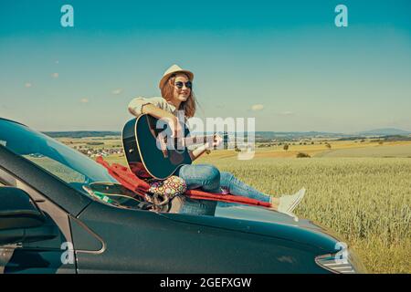 una giovane ragazza che si cimenta con la chitarra sul cofano della sua auto durante il viaggio in campagna Foto Stock