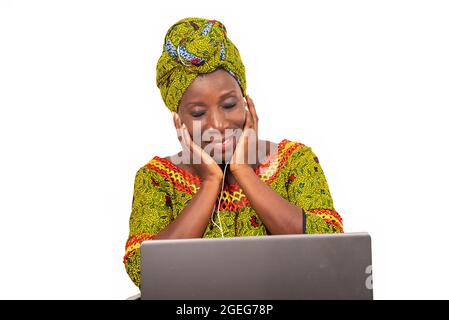 bella donna africana adulta vestita in tessuto di lombo e sciarpa verde, ama ascoltare la musica con le cuffie, nel suo portatile mentre sorride con la sua ey Foto Stock
