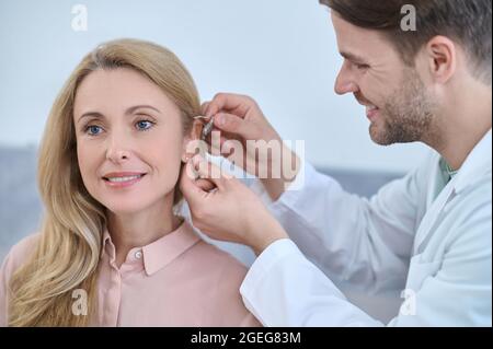 Gioioso medico che fissa un aiuto sordo dietro l'orecchio femminile Foto Stock