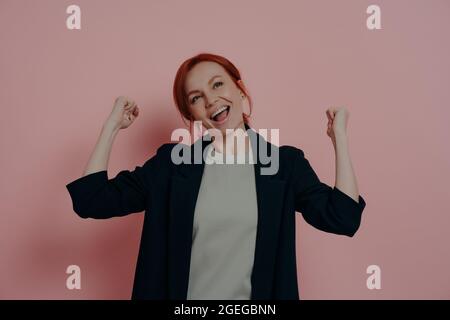 Giovane felice sorgioiata zenzero donna business alzando le mani in pugni e celebrare trionfo Foto Stock
