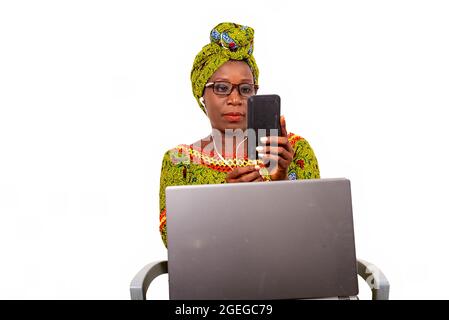 bella donna africana adulta vestita in tela di lombo e sciarpa verde con occhiali, è seduta su una sedia con il suo computer sui suoi piedi, e gode commu Foto Stock