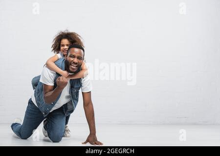 allegra ragazza afroamericana piggybacking padre mostrando vincere il gesto mentre si inginocchiano sul grigio Foto Stock