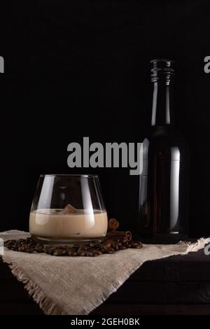 Un bicchiere di liquore al gelato e una bottiglia nera accanto. Sfondo scuro, orientamento verticale. Foto Stock