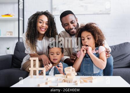 sorprende ragazza afroamericana vicino sorridenti genitori e fratello che giocano a blocchi di legno gioco Foto Stock