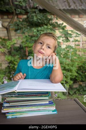 Ragazzo annoiato 5-6 anni seduto a un tavolo di fronte a una pila di libri. Emozione di malcontento, fatica di un preschooler. Sviluppo precoce, apprendimento Foto Stock