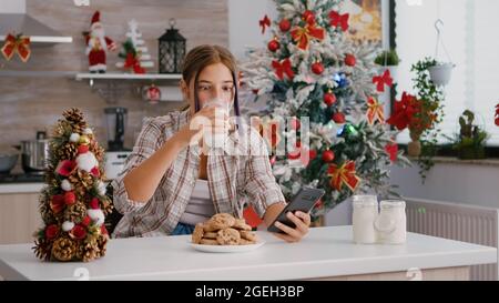 Ritratto di bambini seduti a tavola in una cucina decorata con Natale che naviga sui social media utilizzando lo smartphone. Ragazza caucasica godendo le vacanze di natale mangiando i biscotti fatti in casa tradizionali che bevono latte Foto Stock