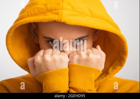 Giovane donna in cappa ocra che tiene i pugni vicino faccia su sfondo bianco. Foto Stock