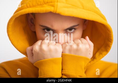 Giovane donna in cappa ocra che tiene i pugni vicino faccia su sfondo bianco. Foto Stock