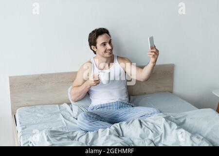 uomo sorridente con una tazza di caffè prendendo selfie sul cellulare a letto Foto Stock