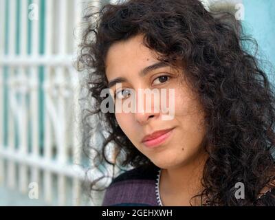 Bella positiva emotiva giovane messicana Latina ispanica business donna con lunghi ricci neri pone di fronte a una griglia di sicurezza bianco finestra in acciaio. Foto Stock