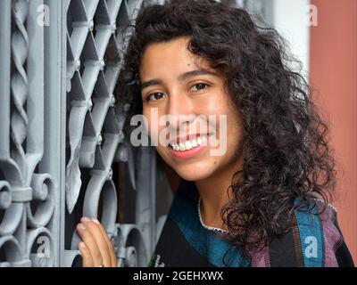 Bella positiva emotiva giovane messicana Latina ispanica donna con lunghi ricci neri pone di fronte a una griglia di sicurezza bianco finestra in acciaio. Foto Stock