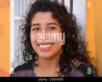 Bella positiva emotiva giovane messicana latina ispanica donna con lunghi ricci neri sorride davanti ad una porta di sicurezza bianca. Foto Stock