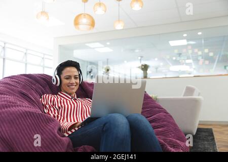 Sorridente donna caucasica per affari creativa indossando le cuffie, sdraiati sul divano e utilizzando il computer portatile Foto Stock