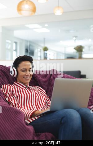 Sorridente donna caucasica per affari creativa indossando le cuffie, sdraiati sul divano e utilizzando il computer portatile Foto Stock