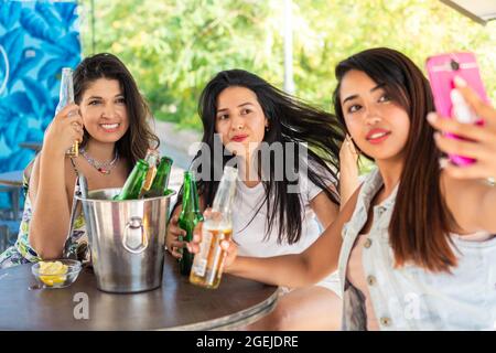 Tre giovani amici latini che hanno la loro foto scattata mentre bevono le birre. Amici delle ragazze che si godono una bella serata estiva su una terrazza. Ragazza amici taki Foto Stock