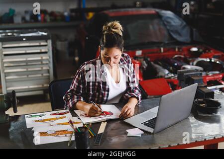 Meccanico misto di auto femminile da corsa seduto alla scrivania, facendo lavoro di documentazione in officina Foto Stock