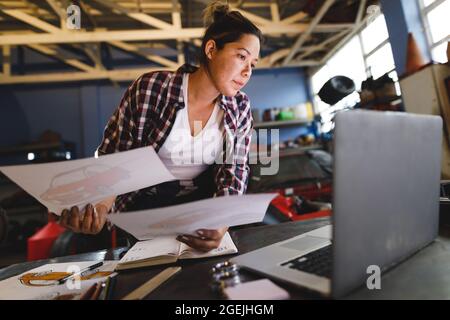 Meccanico misto di auto femminile da corsa seduto alla scrivania, facendo lavoro di documentazione in officina Foto Stock