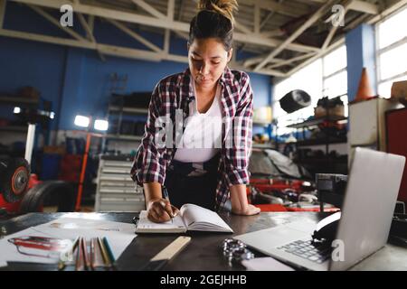 Meccanico misto di auto femminile da corsa seduto alla scrivania, facendo lavoro di documentazione in officina Foto Stock