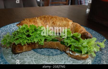 Croissant croccante giallo farcito con insalata, cetrioli su un piatto con motivo e tavolo in legno Foto Stock