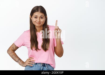 Immagine della giovane donna che mostra il dito sollevato e sorridente, che punta a qualcosa, che insegna lezione, che scuote l'indice, che si alza su sfondo bianco Foto Stock