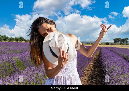 Felice giovane donna che gioca con il suo cappello in un campo di lavanda in fiore. Ha uno sguardo intenso e gli occhi rivolti alla fotocamera. Foto Stock