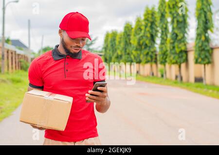 addetto alla consegna africano che controlla il suo telefono mentre trasporta una scatola Foto Stock