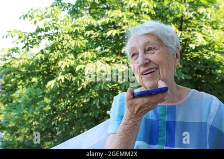 Donna anziana di charme in occhiali rettangolari neri con funzione di riconoscimento vocale sul telefono cellulare. Donna anziana moderna che tiene gadget elettronici, inviando messaggi vocali via messaggero online Foto Stock