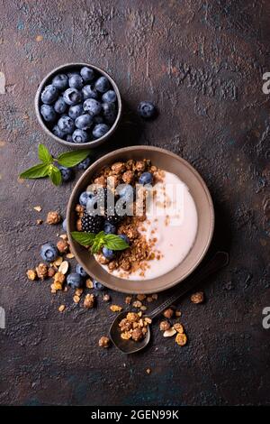 Cibo biologico per una colazione sana e dietetaria. Vista dall'alto della ciotola con granola, yogurt, mirtillo fresco, mora e menta su sfondo di cemento scuro Foto Stock