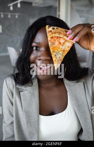 donna afroamericana in tuta d'affari che copre gli occhi con una fetta di pizza Foto Stock