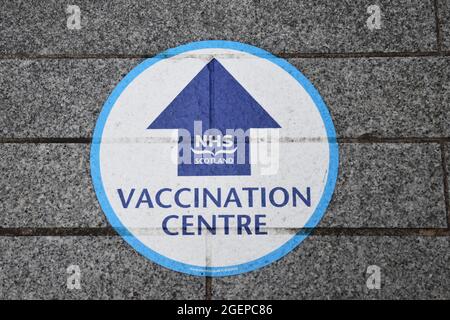 Segno isolato NHS Scotland Vaccine Center in cerchio blu con freccia. Su marciapiede con pietre grigie. Foto Stock