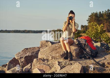 Donna seduta sulle rocce della riva scattando foto con la fotocamera Foto Stock