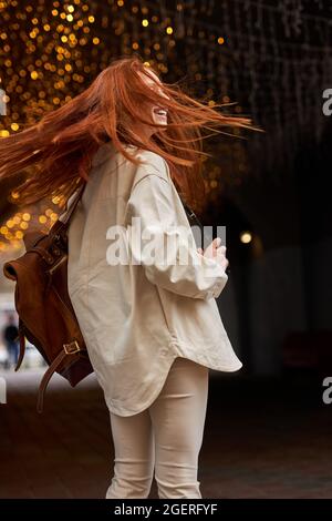 Giovane donna rossa eccitata si girò indietro, con capelli volanti. Foto stile di vita all'aperto. Capelli rossi naturali. La femmina caucasica sta osservando positiva, tenendo re Foto Stock