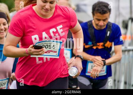 Una donna che indossa una t-shirt rosa controlla il suo telefono mentre corre, mentre un uomo controlla il suo orologio in background, durante un evento Cancer Research Race for Life a Hampstead Heath, Londra Foto Stock