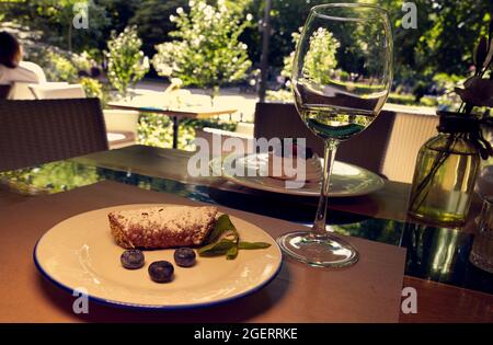 I cannoli siciliani rotolano con mirtilli su un piatto di curcuma bianco e un bicchiere di vino bianco su un tavolo da ristorante con un giardino cittadino sullo sfondo. Foto Stock