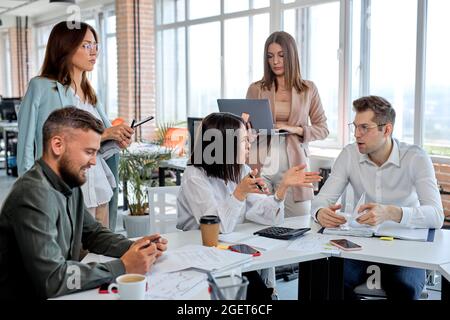 I giovani che lavorano come team in un ufficio moderno, discutono di piani e strategie aziendali, condividono idee. Uomini e donne caucasici in abbigliamento formale con ta Foto Stock