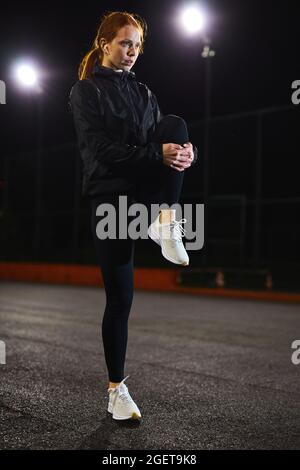 Fit giovane donna caucasica facendo stretching allenamento all'aperto in strada città di notte. Attraente donna focalizzata con capelli rossi naturali impegnati nello sport, Foto Stock