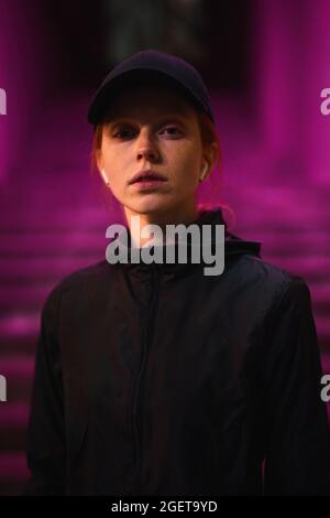 Giovane donna caucasica rossa in berretto nero, abiti sportivi e cuffie che cammina all'aperto solo nella città notturna, posando alla macchina fotografica, la vita notturna concep Foto Stock