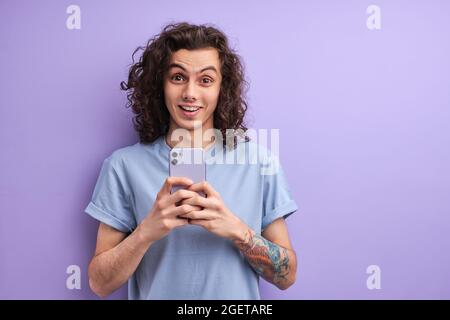 Uomo gioioso con capelli ricci lunghi casualmente vestito tenendo smartphone in mano reagisce emozionalmente sulle notizie mentre legge notizie, navigando in rete Foto Stock