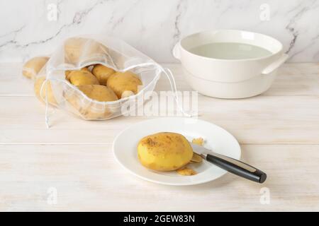 Tuber e coltello in potatoe crudo semisbucciato su un piatto e sacchetto di nylon riutilizzabile con patate biologiche non pelate su tavolo da cucina bianco in legno. Foto Stock