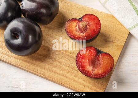 Prugne nere tagliate a metà e intere su un tagliere di legno su un tavolo di legno bianco. Prugne fresche di grandi dimensioni da vicino. Pronto a mangiare frutta e frutti di bosco succosi Foto Stock