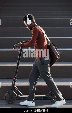 Vista laterale del giovane africano che ascolta la musica in cuffia mentre spinge lo scooter contro la scala Foto Stock