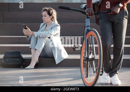 Donna d'affari seria in formalwear testing in smartphone con il giovane uomo che spinge la bicicletta davanti Foto Stock